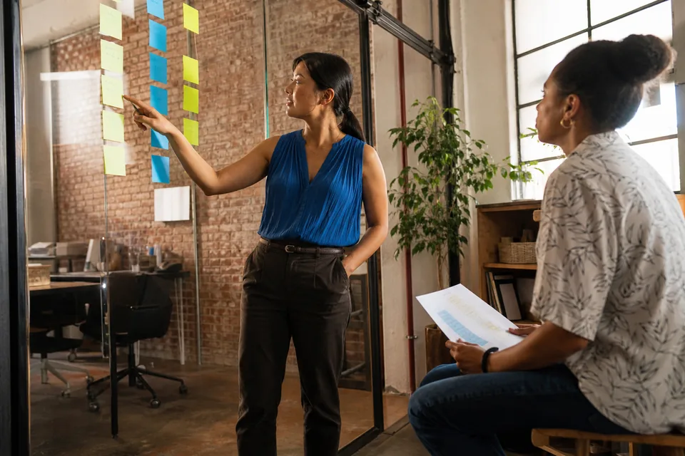 An operations lead sorting BI budget notes into two columns on a glass wall with a colleague